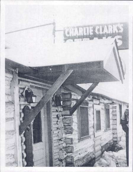Black and white photo of a log building with a canopy and a sign reading "Charlie Clark's Steaks" above the entrance. Snow is visible on the ground.