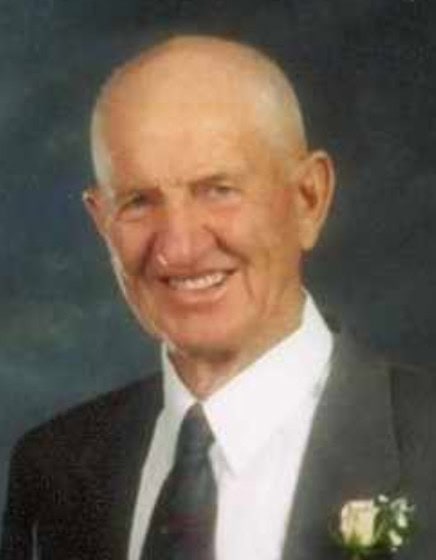 An elderly man with a bald head is wearing a dark suit, white shirt, and tie, with a boutonniere on his lapel, smiling at the camera against a dark background.