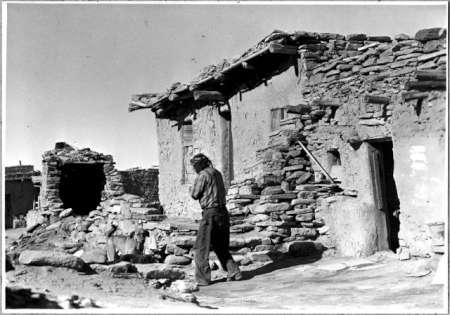 A person stands in front of old, weathered stone and adobe buildings under a clear sky.