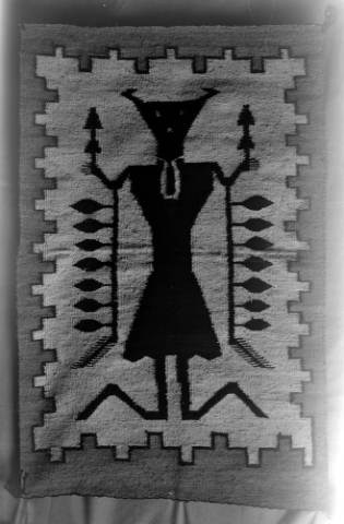 Black and white photo of a woven textile depicting a geometric figure with outstretched arms, symmetrical arrow patterns, and a decorated border.