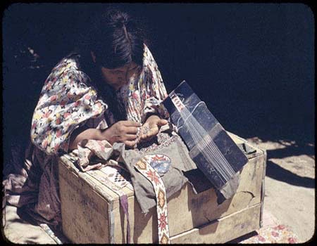 A person wearing a patterned shirt sits at a wooden box, weaving colorful beads with a bead loom.