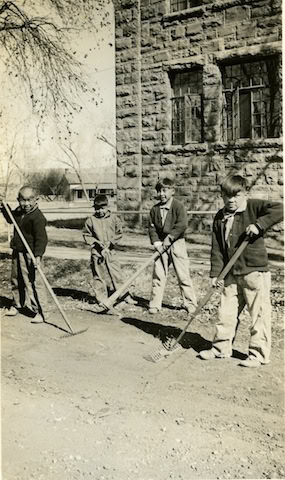Four boys stand outside near a stone building, holding rakes and working on a dirt area. Trees and a house are visible in the background.