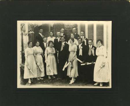 A group of young men and women in formal attire pose outdoors, some holding rolled papers, in front of a clapboard building. The photo appears to be from the early 20th century.