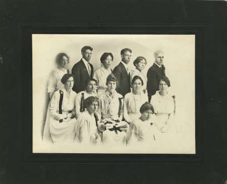 Black and white portrait of thirteen people, eight women seated in front and five individuals, three men and two women, standing behind them, all dressed in early 20th-century attire.