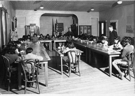 Black-and-white photo of children seated at long tables in a classroom, writing or reading, with two adults standing at opposite ends of the room.