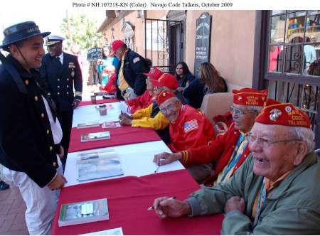 Several elderly men in red uniforms and hats labeled "Navajo Code Talkers" sit at a table, interacting with people at an outdoor event in October 2009.