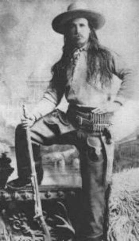 Black-and-white photo of a long-haired person in frontier clothing, wide-brim hat, and gun belt, standing with one foot raised and holding a rifle.