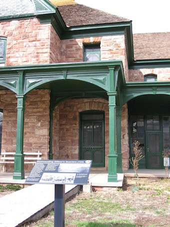 Historic stone house with green wooden porch and informational sign in the foreground.