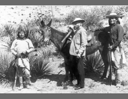 Three men stand in a desert landscape beside a saddled mule; two wear wide-brimmed hats and one holds a rifle. Dry plants are visible in the background.