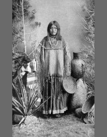 A Native American woman stands in traditional dress, surrounded by pottery, baskets, and desert plants, with a painted shield leaning nearby.