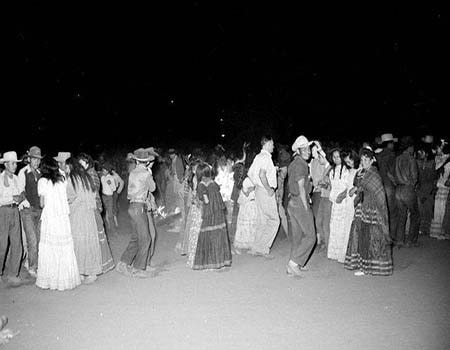 A group of people, many wearing cowboy hats and long dresses, dance together outdoors at night.