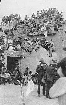 A large group of people, including men, women, and children, gather on and around a multi-level adobe structure, some seated and others standing, in an outdoor setting.