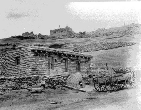 A stone and adobe house stands in a rocky landscape, with a wooden wagon in front and stone ruins visible on the hill behind.