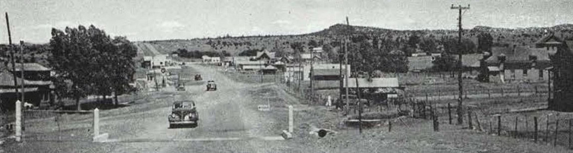 Black-and-white photo of a rural town with cars driving on a wide, unpaved road, lined with houses, utility poles, and scattered trees, with hills in the background.