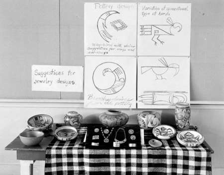 Display of pottery and jewelry on a table with a checkered cloth, in front of posters showing pottery designs and suggestions for jewelry designs.