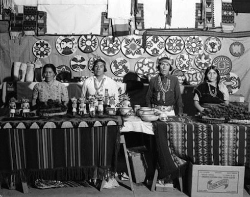 Four women stand behind tables displaying traditional crafts, pottery, and food, with woven textiles and patterned crafts hanging on the wall behind them.