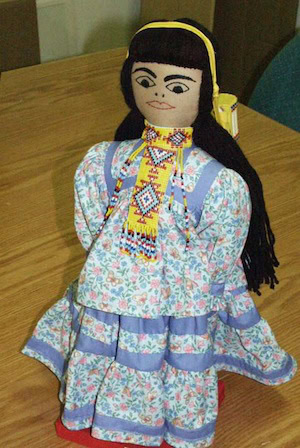 A handmade cloth doll with long black hair, a beaded necklace, and a floral dress with purple trim stands on a wooden table.