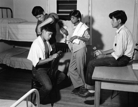 Four young men in a dormitory room; one is getting a haircut while two others observe and one sits on a table holding an object.