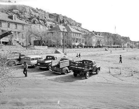 Black and white photo of vintage trucks and cars parked in front of stone buildings with rocky hills in the background; a few people are walking nearby.