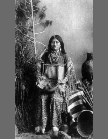 A Native American woman sits in traditional dress, holding a woven basket, surrounded by pottery and a blanket, with plants and a tree in the background.