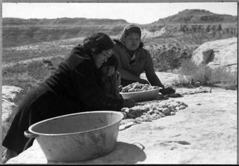 Two women sort and arrange food, possibly potatoes, on a flat rock outdoors with a metal basin nearby and rocky hills in the background.