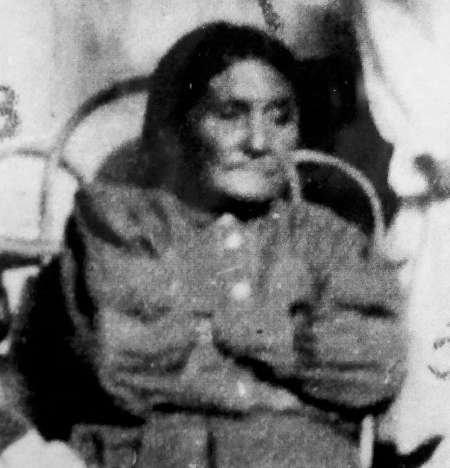 Black and white photo of an older woman with long hair, seated in a wooden chair, wearing a buttoned dress with her arms crossed.