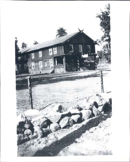 Black-and-white photo of a two-story wooden building with a sloped roof, surrounded by trees and a fence with rocks in the foreground.