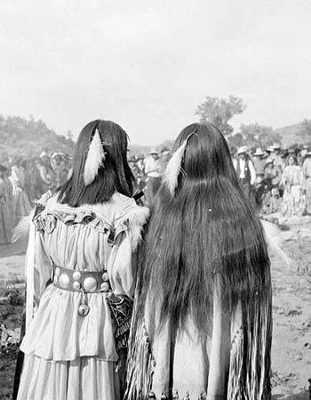 Two people in traditional clothing with feather hair ornaments stand with their backs to the camera, facing a gathered crowd outdoors.