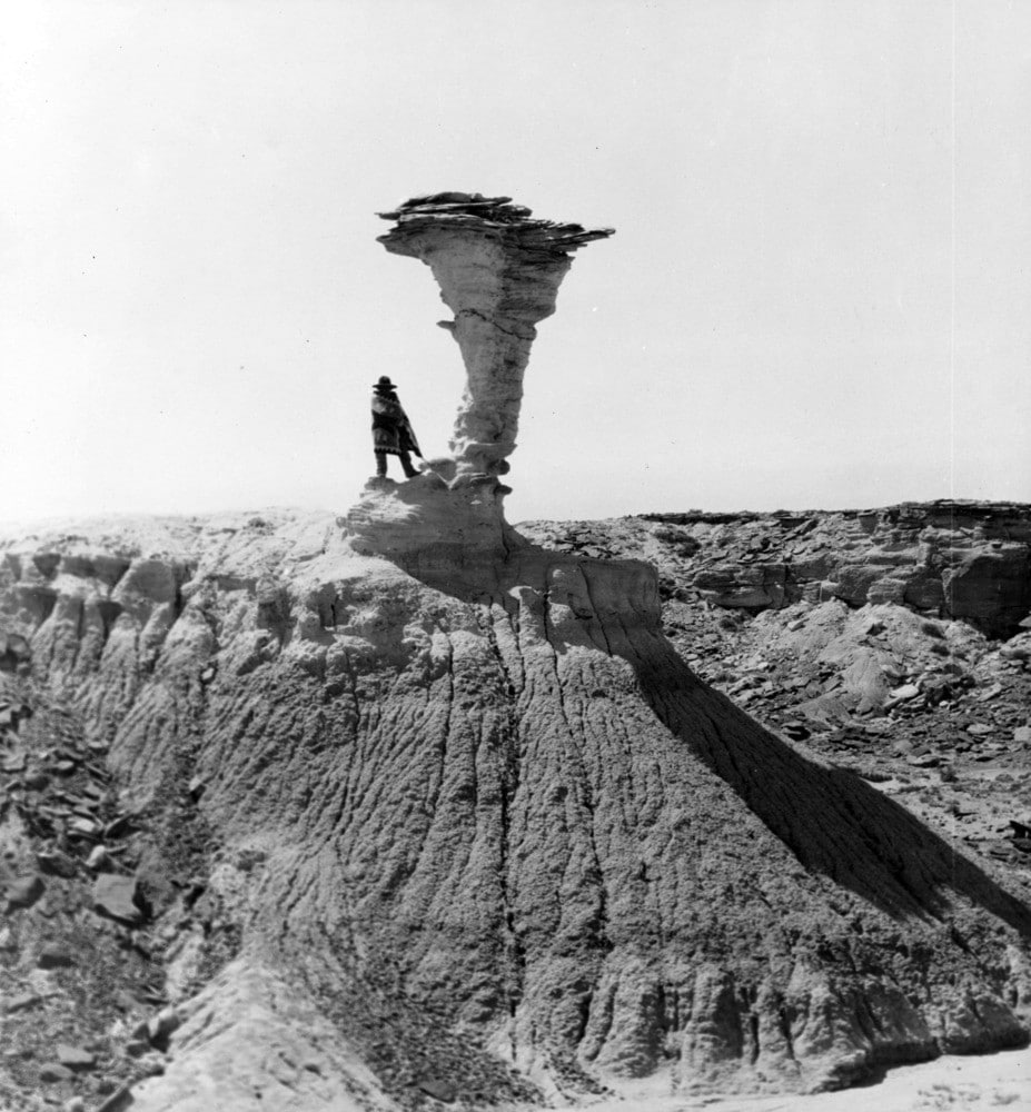 A person stands at the base of a tall, narrow rock formation in a barren, eroded landscape under a clear sky.