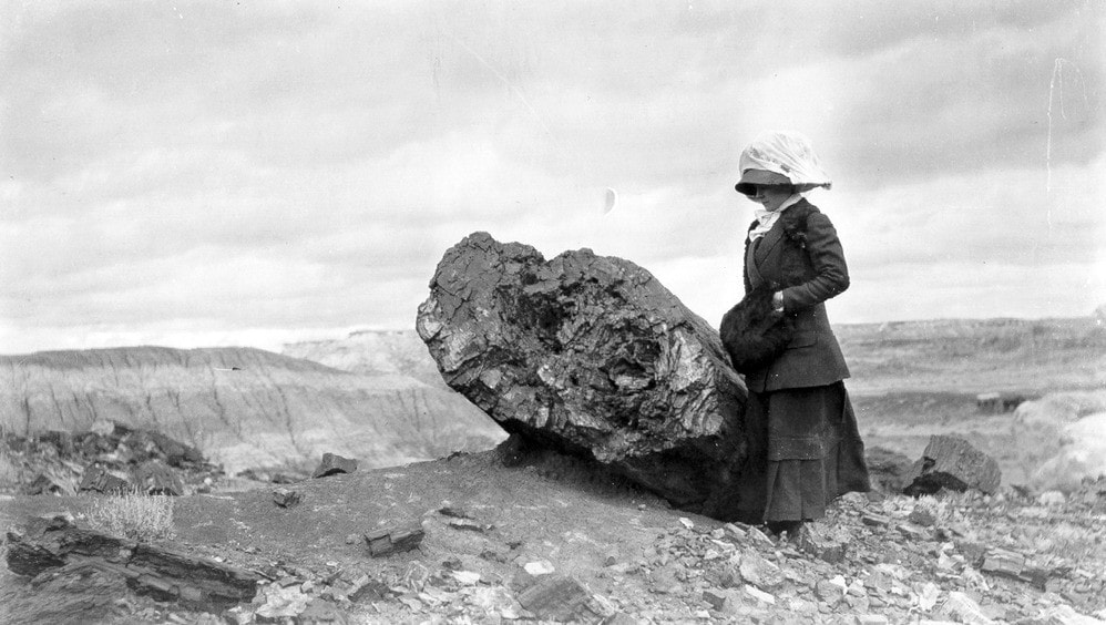 A woman in early 20th-century clothing stands beside a large rock in a barren, rocky landscape under a cloudy sky.