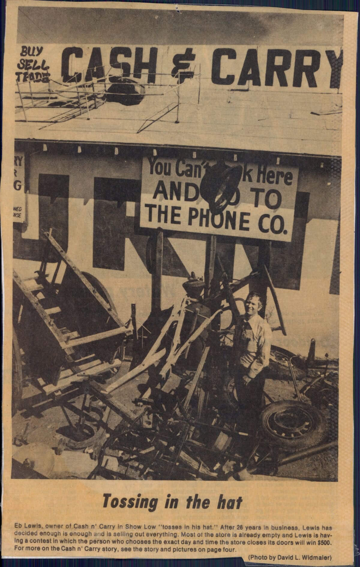 A man stands in front of a Cash & Carry store next to a sign reading "You can look here and go to the phone co." Debris and store items are scattered around him.