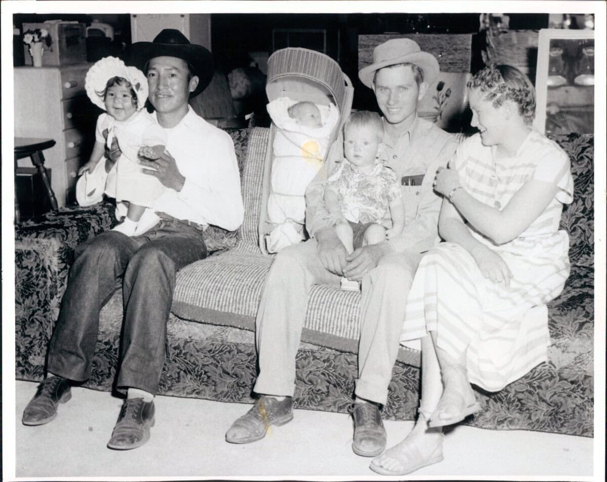 Five people, including two men, a woman, a toddler, and a baby in a stroller, sit together on a patterned couch in a living room, posing for a photo.