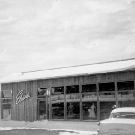 Black-and-white photo of a wooden building with large windows, a sign reading "Elaine," and a vintage car parked in front. Snow is visible on the ground and roof.