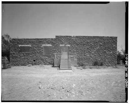 A rectangular stone building with three shuttered windows and a central staircase leading to a closed door, set against a clear sky and barren ground.