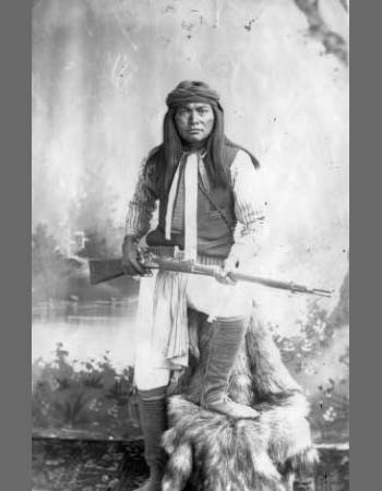 A Native American man wearing traditional clothing, seated on a fur-covered object, holding a rifle, with a painted backdrop behind him.