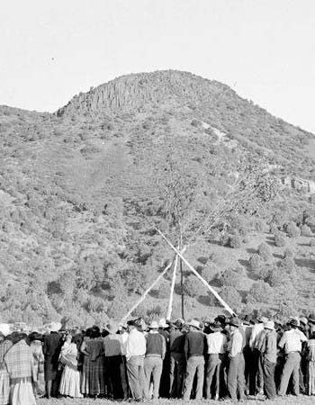 A group of people stands in a circle outdoors near a tall wooden pole structure, with a hill covered in vegetation in the background.