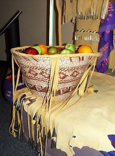 Woven basket with a geometric pattern filled with various fruits, resting on a fringed, beaded surface with leather and fabric.