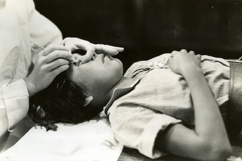 A person lies on their back while another person, wearing a glove, examines or treats their eye.
