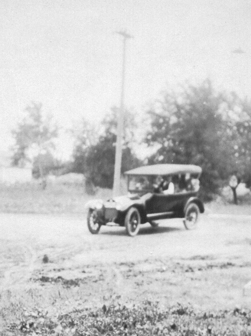 A black and white photo of an early 20th-century car with a soft top driving on a dirt road, trees and utility pole in the background.