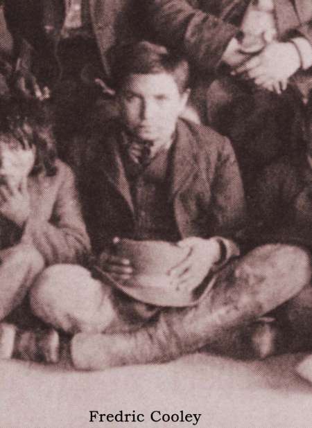 A young boy sits cross-legged on the floor, holding a hat in his lap, with other children partially visible beside him.