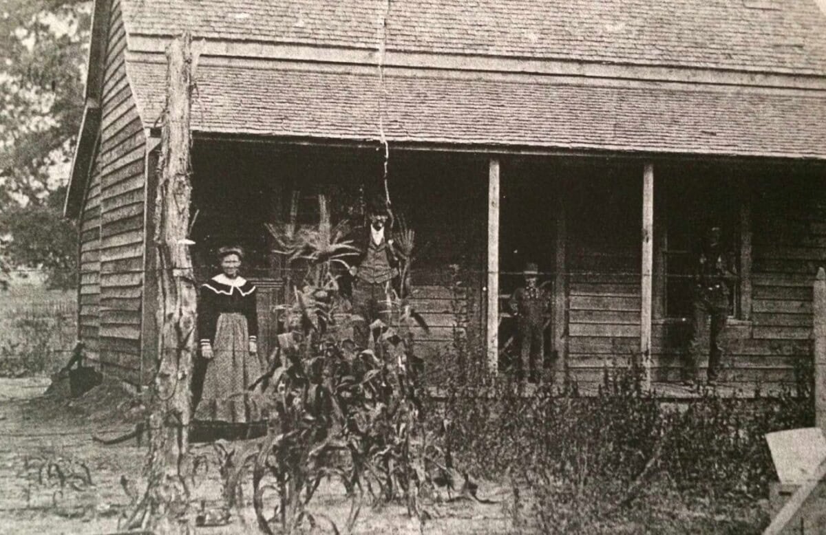 Black and white photo of four people standing outside a wooden house; one woman is near the corner, three others are on the porch, with tall plants in the foreground.