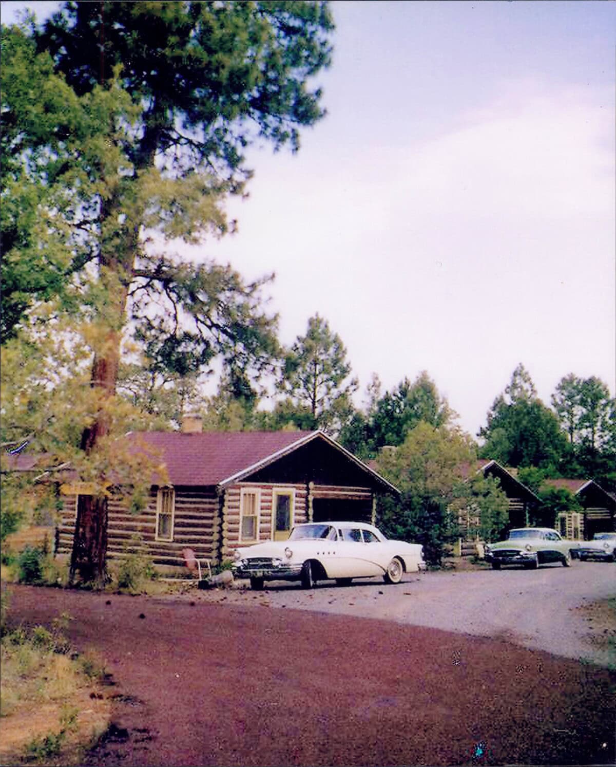 A vintage photo of log cabins surrounded by trees, with a white classic car and another vehicle parked on a paved area in front.