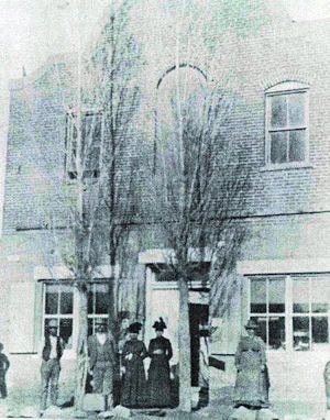 Black and white photo of several people standing in front of a two-story brick building with arched and rectangular windows and two trees by the entrance.