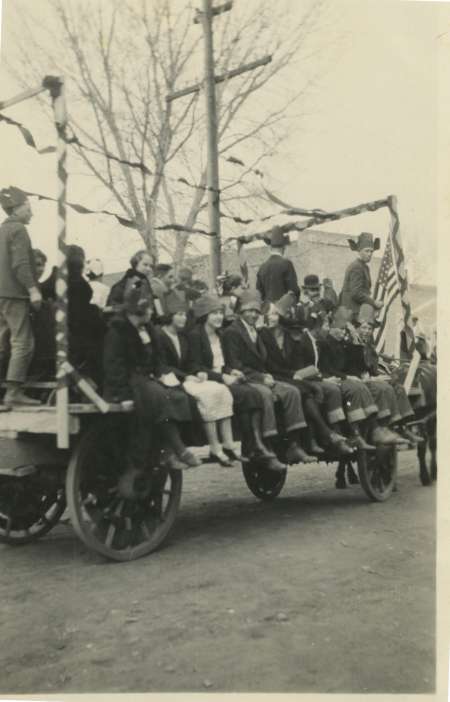 A group of people sit on a decorated horse-drawn wagon, dressed in coats and hats, with leafless trees and a utility pole in the background.