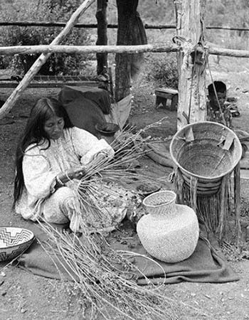 A woman sits on a blanket outdoors, weaving baskets from natural materials, with completed baskets beside her under a simple wooden shelter.