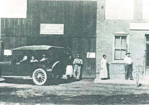 A vintage photo shows a car with several people inside parked outside a brick building with a “Ford” sign; three people stand nearby on the dirt ground.