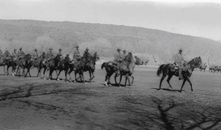 A group of mounted soldiers rides in formation across a flat, open landscape with hills in the background.