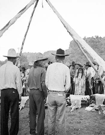 A group of people, some in hats and traditional clothing, gather outdoors under a tipi frame with animal hides on the ground.