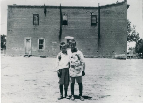 Two young boys stand together in front of a large, plain brick building on a dirt lot in an old black and white photograph.