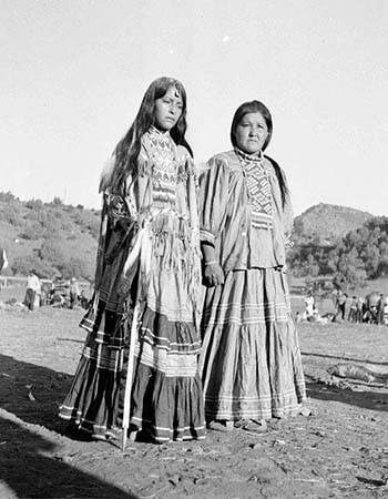 Two Indigenous women wearing traditional dresses stand outdoors on dirt ground, with hills and trees visible in the background.